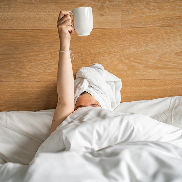 Frau im Bett mit Handtuch auf dem Kopf die eine Kaffeetasse in die Luft hält